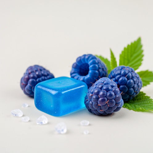 Morceaux de chewing-gum bleu brillants disposés avec des framboises bleues et des feuilles vertes sur une surface claire.